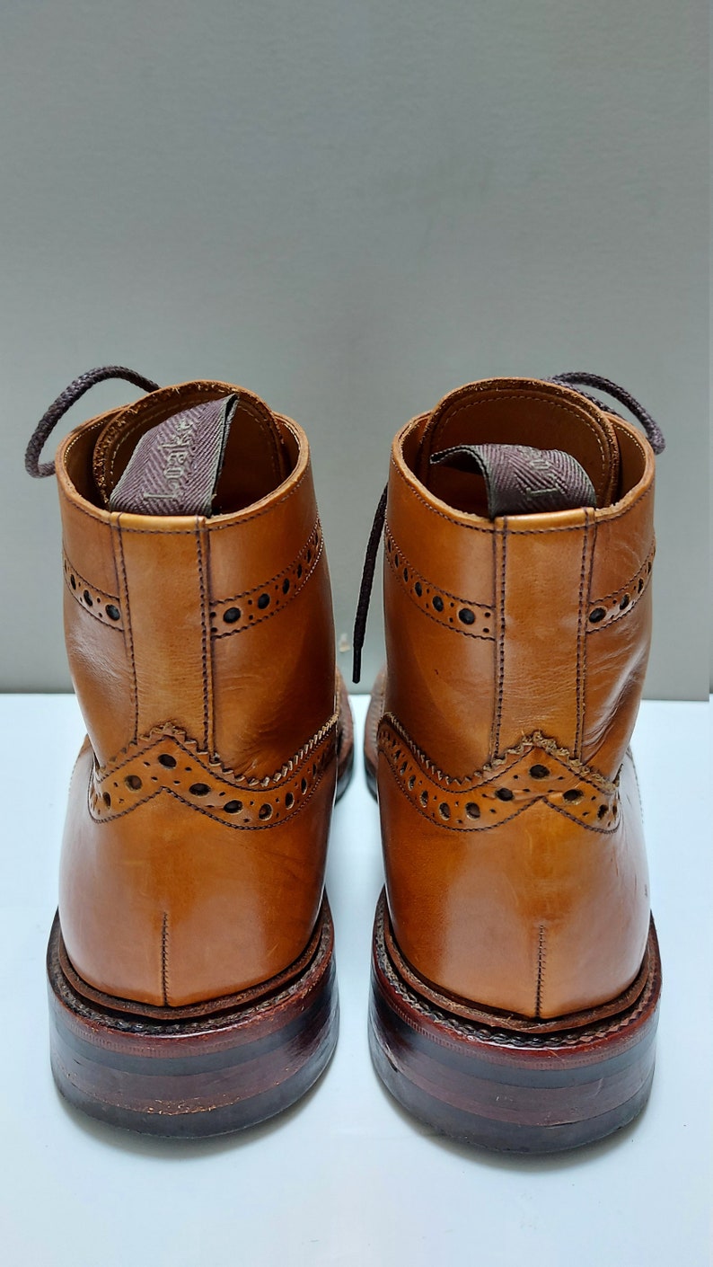 May include: A pair of brown leather lace-up boots with brogue detailing. The boots have a brown leather sole and a brown leather heel.