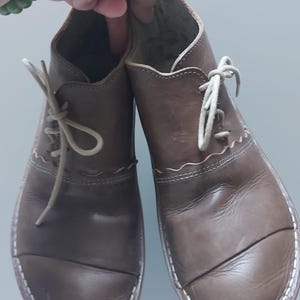 以下が含まれることがあります： 縁に白いステッチが施された、茶色の革製のレースアップシューズのペア。靴は丸みを帯びたつま先とわずかに盛り上がったヒールを持っています。