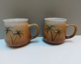 May include: Two brown ceramic mugs with a white glaze and a palm tree design. The mugs have a rounded shape and a handle.