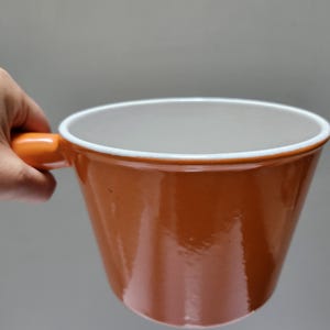 May include: An orange ceramic mug with a white interior and a matching orange handle. The mug has a glossy finish and a simple, modern design, suitable for coffee or tea.