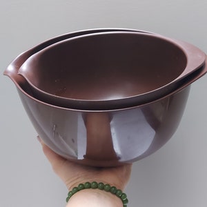May include: Two brown mixing bowls, one inside the other. The bowls have a rounded shape with a flared rim and a handle-like extension on one side. The bowls are made of a smooth, glossy material.
