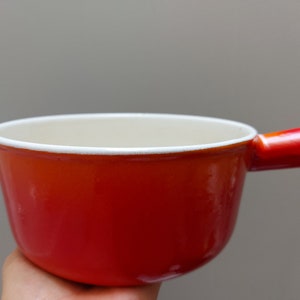 May include: A red and orange enamel cast iron pot with a white rim and a single handle. The pot is empty and appears to be new.