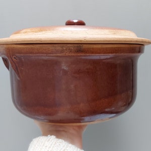 Puede incluir: Una olla de cerámica marrón con una tapa de madera. La olla tiene una forma redondeada y un acabado liso y brillante. La tapa tiene un pequeño pomo en la parte superior.