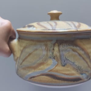 Könnte beinhalten: Eine Keramik-Auflaufform mit Deckel, die ein braunes, gelbes und graues Wirbelmuster aufweist. Die Form hat zwei Griffe an den Seiten.