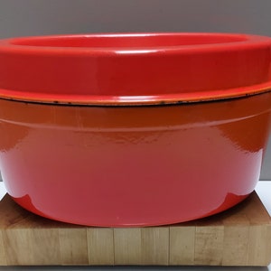 Puede incluir: Cazuela de hierro fundido esmaltado naranja con tapa. La olla está sobre una tabla de cortar de madera.