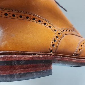 May include: Close-up of the sole of a brown leather brogue boot with a brown leather welt and a black rubber sole.
