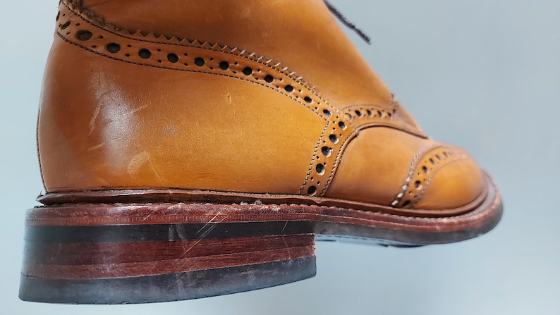 May include: Close-up of the sole of a brown leather brogue boot with a brown leather welt and a black rubber sole.
