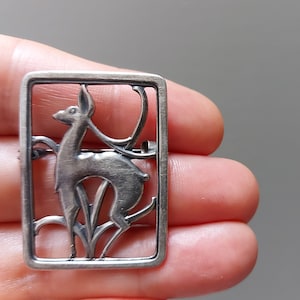 May include: A silver brooch featuring a stylised deer in a rectangular frame. The deer is depicted in a standing position with its head turned to the side. The brooch is a delicate and intricate piece of jewellery.