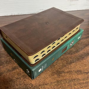 May include: A brown leather-bound Bible with gold-edged pages rests atop a green book. The brown cover has a small emblem in the center. The pages are tabbed for easy reference. The books are on a wooden surface.