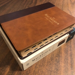 May include: A brown leather-bound Bible with the title "MacArthur Study Bible" embossed in gold lettering. The Bible is closed and sitting on a white box with the same title.