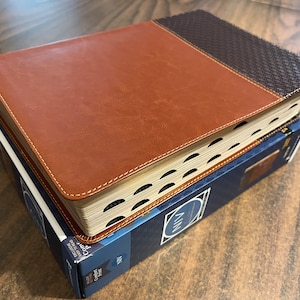May include: A brown leather-bound book with a black woven pattern on the spine. The book is closed and sitting on top of a blue box with the word "NIV" printed on it.