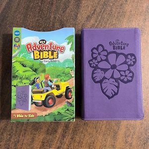 May include: Two purple hardcover Bibles with the text "NIrV Adventure Bible" on the front cover. The left Bible is for early readers and has a colorful illustration of a jeep driving through a jungle. The right Bible has a floral design embossed on the cover.