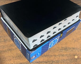 Op de afbeelding: Een zwart lederen gebonden boek met een zilveren rug en een blauwe en zilveren doos met het woord "NIV" erop.