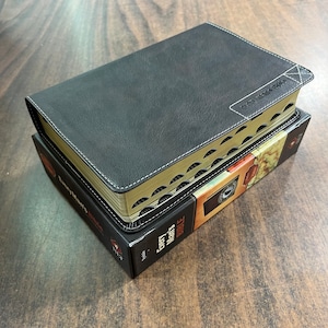 May include: A black leather-bound Bible with gold-gilded edges and white stitching. The book is resting on its original box, which features a map design and the text "Every Man's Bible".