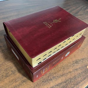 May include: A burgundy leather-bound Bible with gold-gilded edges, resting on a matching box. The cover features the words "SANTA BIBLIA" and a gold olive branch design. The book is closed, with a ribbon bookmark.