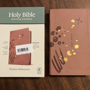 May include: A pink leather-bound Holy Bible with gold accents and a dragonfly design. The Bible is a Thinline Reference edition and is published by New Living Translation.