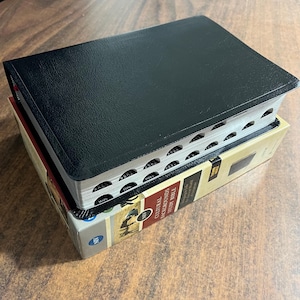 May include: A black Bible with a textured cover rests atop a box labeled "Cultural Backgrounds Study Bible." The Bible's pages have labeled tabs. The box is beige, red, and brown with text and illustrations. The image is shot from a high angle.