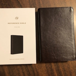 May include: A Reference Bible with a dark brown leather cover next to a white book cover. The white cover has the text "REFERENCE BIBLE" and "ENGLISH STANDARD VERSION". The Bible is a coffee-colored edition.