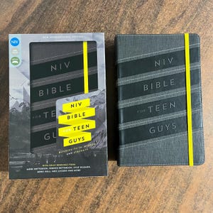 May include: A gray Bible for Teen Guys with the text "NIV BIBLE FOR TEEN GUYS" and a yellow elastic band. The book is next to its box, which also has the text "NIV BIBLE FOR TEEN GUYS" and a mountain graphic.