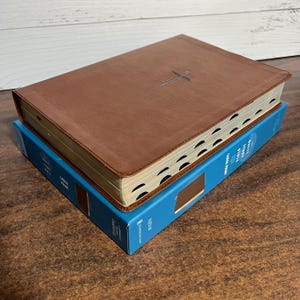 May include: A brown leather-bound Bible with a cross design is stacked on top of a blue box. The Bible has gold-edged pages and a stitched cross on the cover. The blue box has white text and a brown image of a book.