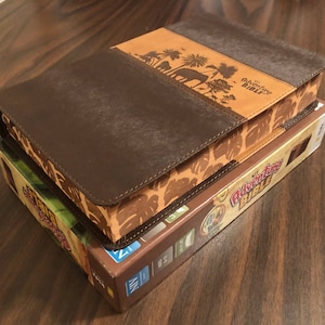 May include: A brown leather-bound book with a tan cover featuring a jungle scene with a large elephant. The book is titled "The Adventure Bible".