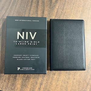May include: A black NIV Thinline Bible with large print and a matching black leather cover. The book's cover displays the text "NIV" in large white letters, along with details about the typeface, leather, and text color. The Premier Collection logo is also visible.