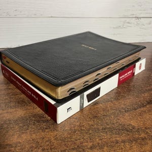 May include: A black leather-bound Holy Bible with gold-gilded edges rests atop a red and white box labeled "Giant Print Thinline Bible." The book has a textured cover and the words "Holy Bible" are embossed in gold.