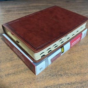 May include: Two books stacked on a wooden surface. The top book is a brown leather-bound Bible with gold-colored edges and a decorative border. The bottom book is a white and red book with the text "CENTER-COLUMN REFERENCE BIBLE".