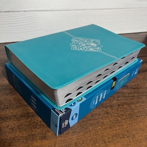 May include: A teal blue leather-bound Bible with a floral design on the cover. The Bible is closed and sitting on top of a blue box.