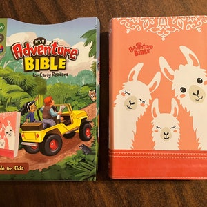 May include: Two children's Bibles. The first is a paperback with a colorful illustration of a jeep driving through a jungle. The text on the cover reads "NIRV Adventure Bible for Early Readers." The second is a hardcover with a pink cover and three white llamas with closed eyes. The text on the cover reads "The Adventure Bible."