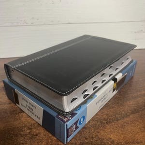 May include: A black leather-bound Bible with silver accents and a white stitched border. The Bible is closed and sitting on a blue and white box with the text "Personal Size Bible Large Print" on the side.