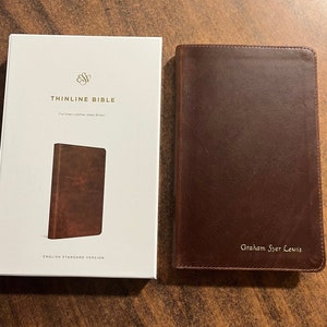 May include: A brown leather-bound Bible with the words "THINLINE BIBLE" and "ENGLISH STANDARD VERSION" printed on the front cover. The Bible is closed and has a personalized inscription that reads "Graham Syer Lewis".