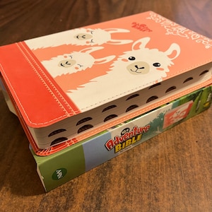 May include: A pink and white children's Bible with a cover featuring three llamas. The title "My Adventure Bible" is printed on the spine.