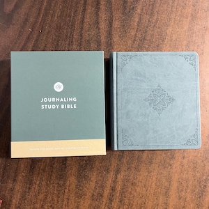 May include: A teal-colored "Journaling Study Bible" with a matching box. The box has the title in white text and a logo. The Bible has an embossed design on the cover. The box and Bible are on a wooden surface.