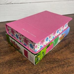 May include: A pink hardcover children's Bible with a floral design on the spine. The Bible is personalized with the name "Millie Rose Lawson".