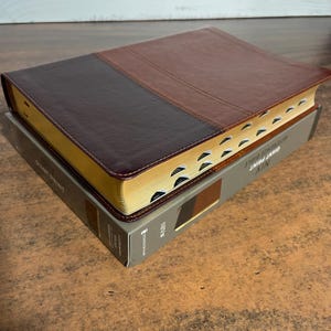 May include: A two-tone brown leather-bound Bible with gold-gilded edges, resting on a gray box. The Bible has a dark brown top and a lighter brown bottom. The box reads "NIV Giant Print Reference Bible."