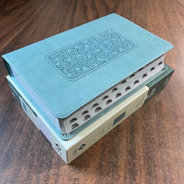 May include: A light blue hardcover Bible with a decorative floral design on the front cover. The Bible is closed and sitting on a wooden surface.