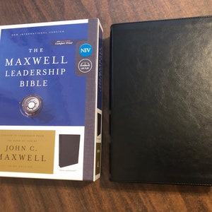 May include: The Maxwell Leadership Bible, New International Version, NIV, with a black leather cover and a blue and gold dust jacket. The dust jacket features the title "The Maxwell Leadership Bible" and the author "John C. Maxwell".
