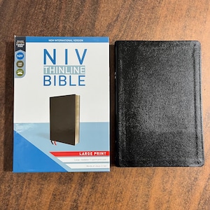 May include: A black Bible with a matching black cover next to a book with a white and blue cover. The cover of the book on the left reads "NIV THINLINE BIBLE" in large print. The book on the right is closed.
