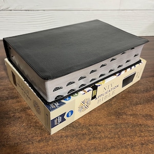 May include: A black leather-bound NIV Application Bible with thumb index tabs, resting on its original box. The box is beige with blue and gold accents, and the text "NIV Application Bible" is visible. The Bible is a religious text.