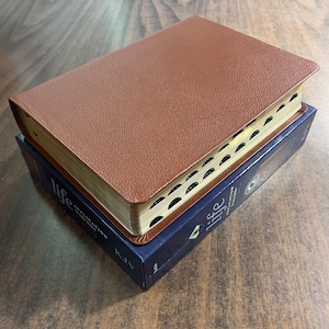May include: A brown leather-bound Bible with gold-edged pages rests atop a blue box. The Bible has black tabbed page markers. The box has the word "life" printed on it.