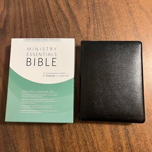 May include: A black leather-bound Bible with the title "Ministry Essentials Bible" on the cover. The cover also includes the text "A Comprehensive Bible for Everyone in Leadership". The book is displayed next to its box, which features a green and white design.