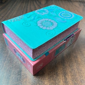 May include: A teal blue hardcover journal with a floral design on the cover. The journal is closed and sitting on top of a pink box with a floral design.