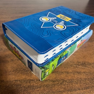 May include: A blue hardcover book with the title "Adventure Bible" on the cover. The cover features a star with binoculars inside and a blue and white topographic map design.