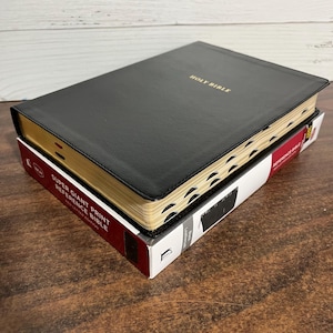 May include: A black leather-bound Holy Bible with gold-gilded edges and the title "HOLY BIBLE" embossed on the front cover. The Bible is in a box with the text "SUPER GIANT PRINT REFERENCE BIBLE NEW LEATHER EDITION" printed on the side.