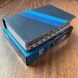 May include: A navy blue Bible with two blue stripes across the cover, resting on a blue box. The Bible has silver-edged pages and a sewn binding. The box reads "BOYS LIFE APPLICATION STUDY BIBLE".