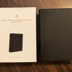 May include: A white box with the text "Single Column Journaling Bible Large Print" and a black leather-bound journal. The journal is closed and has a gold embossed title on the spine.