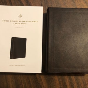 May include: A brown leather-bound journal with the text "Single Column Journaling Bible Large Print" on the cover. The journal is closed and sitting on a wooden surface.