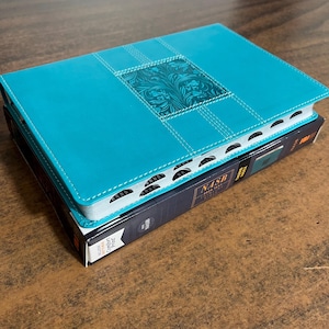 May include: A teal blue leather-bound Bible with a decorative floral design on the front cover. The Bible is closed and the spine is visible. The text "NASB" is printed on the spine.