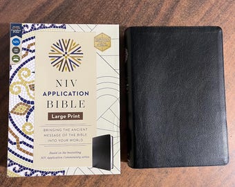 Op de afbeelding: Een zwarte, leren gebonden NIV Application Bible met grote druk. De omslag heeft de titel "NIV Application Bible" en "Grote Druk" in gouden letters. Het boek wordt naast zijn doos getoond, die een geometrisch patroon en het NIV-logo heeft.